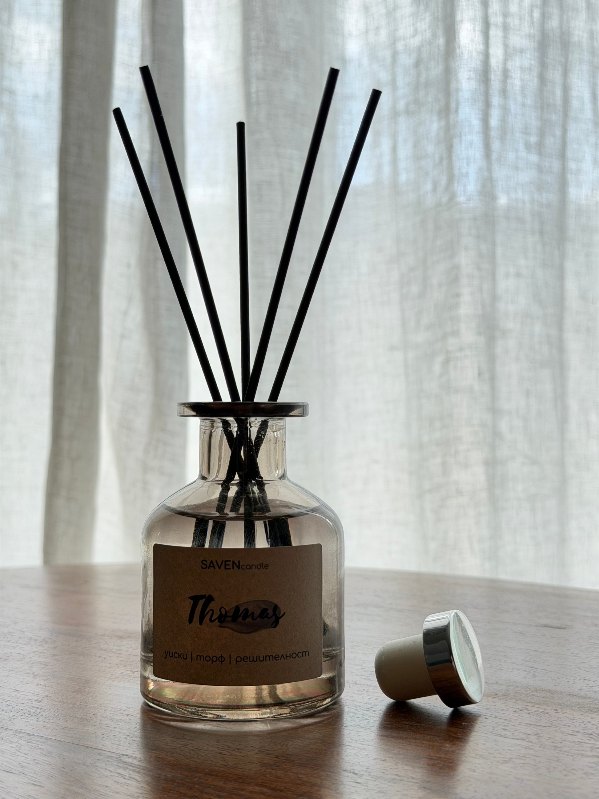 A reed diffuser bottle with black reeds on a wooden table in front of sheer white curtains. The bottle has a clear glass body, a beige label reading 'SAVENcandle' and 'Thomas', and a small beige-and-silver cap lying beside it.