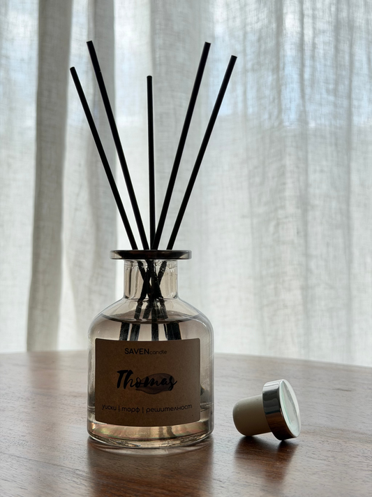 A reed diffuser bottle with black reeds on a wooden table in front of sheer white curtains. The bottle has a clear glass body, a beige label reading 'SAVENcandle' and 'Thomas', and a small beige-and-silver cap lying beside it.